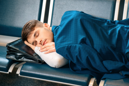 Tired and overworked. Exhausted business man in formalwear sleeping on the rows of chairs in the hall of the airport covering his shoulders with a jacketの写真素材