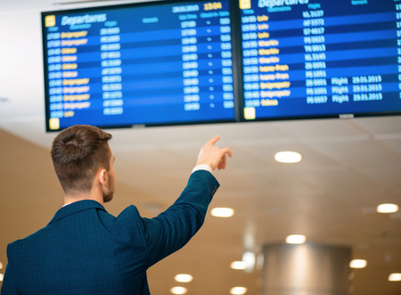 Checking schedule details. Rear view of handsome businessman in formalwear checking time and pointing at flights schedule while standing in the airportの写真素材
