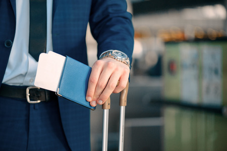 Successful check-in. Cropped image of a handsome young businessman in suit looking at his watch and holding his passport with a ticket while standing in the airport with his luggageの写真素材