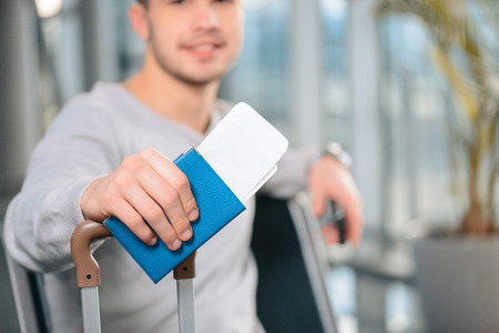 Ready for the flight. Closeup image of passport and luggage of a handsome smiling man in casual wear sitting in the hall of the airportの写真素材