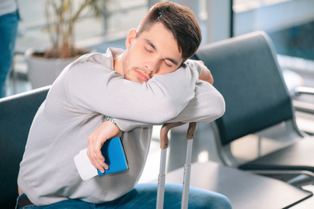Exhausted traveler. Tired handsome man in casual clothing sleeping on his luggage while sitting on the rows of chairs in the airport lobby with a ticket and passport in handの写真素材