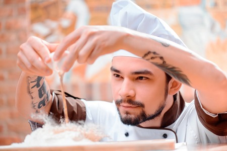 Preparing dough for pastry. Closeup image of handsome inspired cook in uniform and hat cracking eggs upon flour on wooden cutting board in bakers kitchenの写真素材