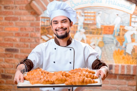 Confident baker at work. Cheerful young baker in apron showing a tray of fresh baked croissants and...