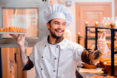 Confident baker at work. Cheerful young baker in apron putting g a tray of fresh baked croissants out of the oven and showing his thumb up while standing on the background of the bakery shopの写真素材