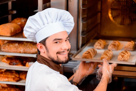 Fresh baked bread. Confident young man in apron taking the fresh baked bread from oven and looking over his shoulder with a smile in the bakery shopの写真素材