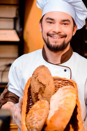 Proud of his baked goods. Cropped image of a handsome young man in apron holding basket with baked goods in bakery shopの写真素材