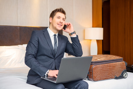 Staying connected. Confident young businessman in suit and tie working on laptop while sitting on the bed in hotel room and talking over his mobile phoneの写真素材
