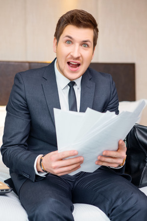 Emotional businessman. Confident young businessman in suit and tie reading documents and looking shocked while sitting on the bed in luxury hotel roomの写真素材