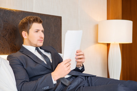 He likes business trips. Confident young businessman in suit and tie reading documents while sitting on the bed in luxury hotel roomの写真素材