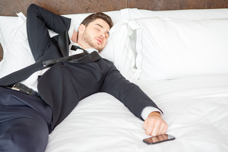 Tired and overworked. Exhausted business man in formalwear sleeping on the bed of the luxury hotel room keeping his hand on mobile phoneの写真素材