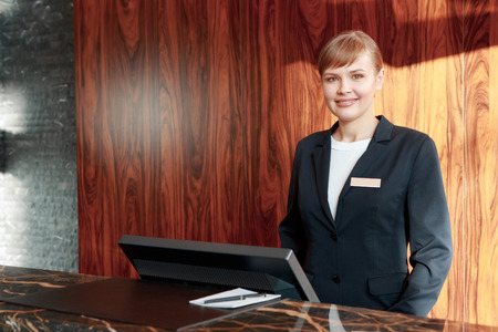 Beautiful stylish hotel receptionist standing behind the service desk in a hotel lobby looking at a...