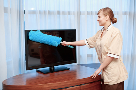 No trace of dust. Hotel maid in beige uniform dusting with a feather duster TV screen in hotel roomの写真素材