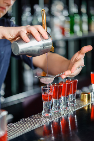 Mixing cocktails. Close-up of barman with cocktail shaker pouring red alcoholic drink into glasses in barの写真素材