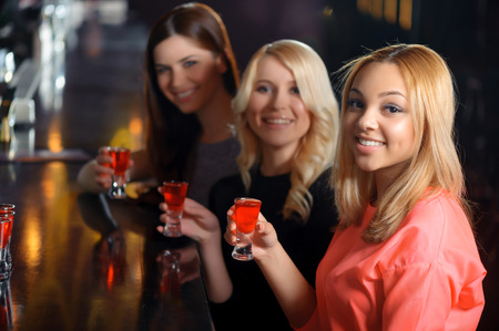 Tasty cocktails. Three beautiful girls holding shots at the bar counter and smiling at a cameraの写真素材