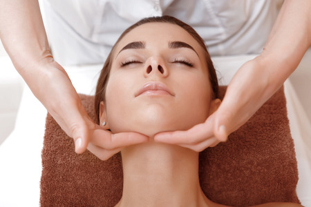 Face lifting massage. Close-up of a masseuse hands massaging face of a young female client of a spa salonの写真素材