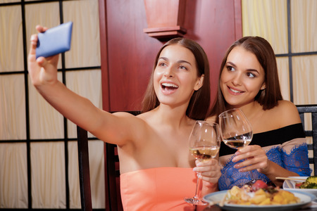 Beauty queens. Two cheerful smiling young women doing selfie with a smartphone in Asian restaurantの写真素材