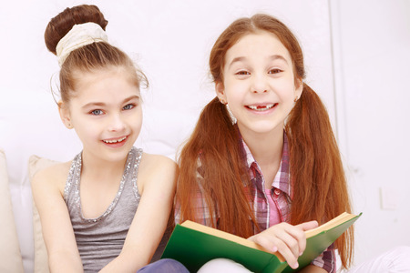 Smile is all you need. Two cute little girls sitting reading book and frankly smiling.の写真素材