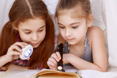 At any time of day. Two nice little girls lying covered with blanket and reading book by using lanternの写真素材