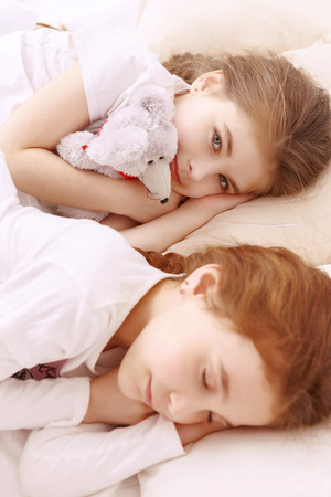 Rest-hour. Two little cute girls lying next to each other on white bed and sleeping with teddy bear toyの写真素材
