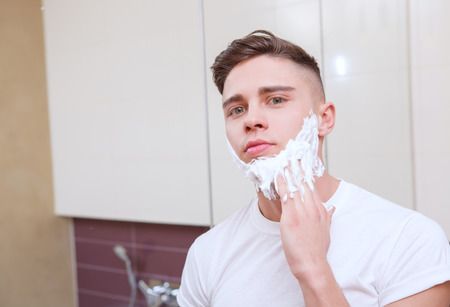 Feeling smooth. Youthful man standing in front of mirror and smoothing out shaving gel.
