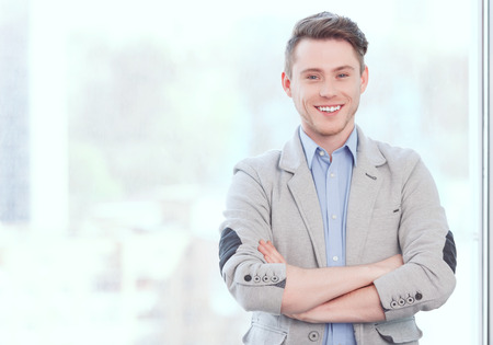 Stay positive. Portrait of young smiling businessman standing in front of window and folding his arms.の写真素材