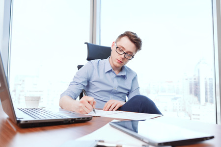 Attentive writing. Young handsome businessman sitting at desk covered with notebook papers and...