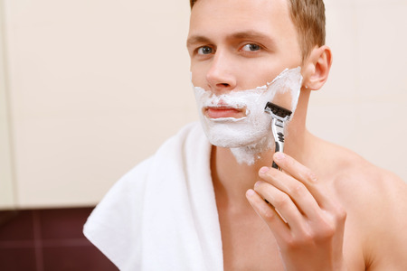 Shaving clean. Handsome topless man shaving in front of mirror and holding towel on his shoulder.