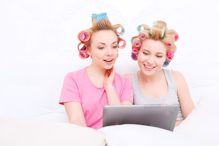 Slumber party. Two young beautiful blond girls wearing pajamas and colorful hair rollers sitting in white bed and looking at the tablet smilingの写真素材