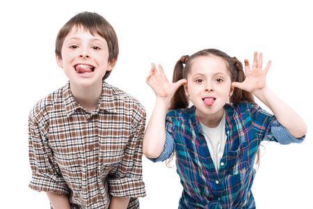 Small pretty girl showing tongue and playfully holding her hands near her head while her older brother showing tongue isolated on white backgroundの写真素材