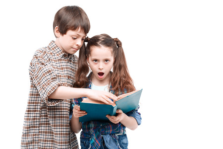Small pretty girl holding a big blue book and looking at you with her mouth wide opened and a surprised face while her older brother, looking at the book isolated on white backgroundの写真素材