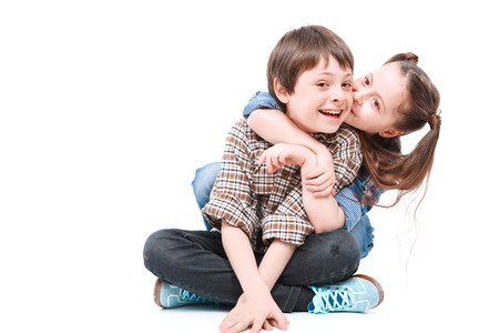 Happy siblings. Portrait of a small handsome boy sitting on the floor with his legs crossed and his small sister sitting behind him hugging and kissing his cheek isolated on white backgroundの写真素材