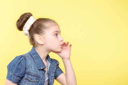 Portrait of a small pretty girl standing sideways and calling someone holding her hand near her mouth wearing a grey t-shirt and jeans jerkin ,isolated on a yellow backgroundの写真素材