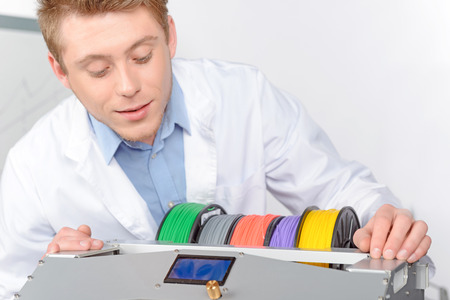 Young handsome scientist checking carefully the colors of a 3D-printer before making a model, wearing white lab coat in laboratoryの写真素材