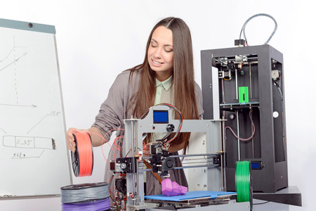 Beautiful young brunette student with long hair holding colorful plastic parts of a 3d-printer...
