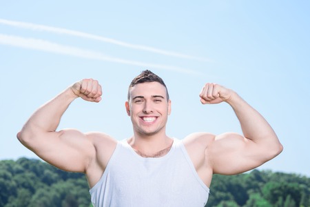 Portrait of a young handsome muscular sportsman posing holding his hands above him, showing biceps and smiling, wearing white sportswear making body exercises full length in the parkの写真素材