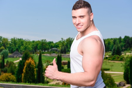 Portrait of a young handsome muscular sportsman posing showing thumb up, wearing black white sportswear during training in the parkの写真素材