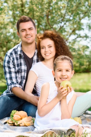 Small pretty girl sitting in the foreground on a plaid smiling and holding a big green apple while her happy parents on a background hugging and smiling, selective focusの写真素材