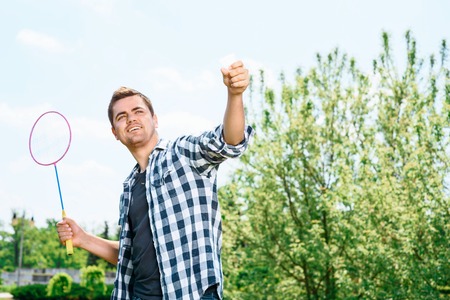 Portrait of a young handsome guy standing in a park holding a racquet above him and a shuttlecock in another hand, ready for making a serveの写真素材