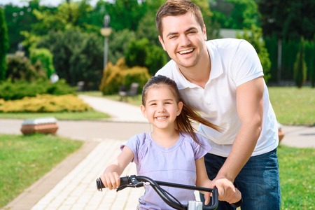 Portrait of a happy caring father teaching his small pretty daughter riding a bicycle in a green park ,smiling waist up photoの写真素材