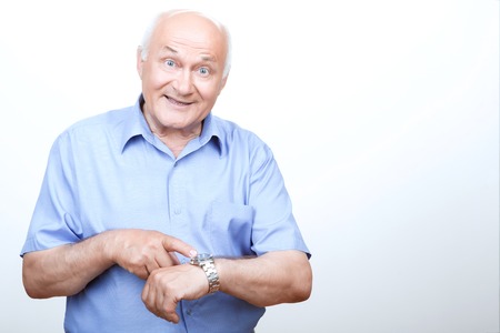 It is high time. Smiling grandfather pointing his wristwatch and looking ahead while standing isolated on white background.の写真素材