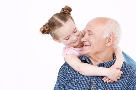 Portrait of a grandfather wearing blue checkered shirt sitting and small pretty granddaughter standing behind him smiling and hugging, looking at each other, isolated on a white backgroundの写真素材