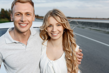 Car trip. Close up portrait of a handsome young man and beautiful blond girlfriend with curvy hair standing hugging near their white car, stunning landscape on the backgroundの写真素材