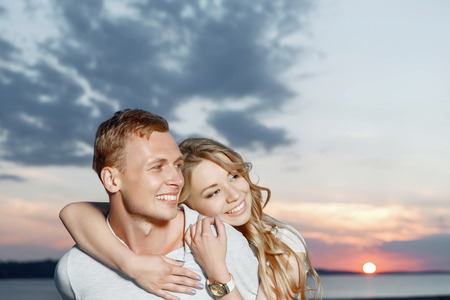 Sweet couple. Portrait of a young beautiful blond woman with curvy hair hugging her handsome boyfriend smiling, stunning sky and sunset on the backgroundの写真素材