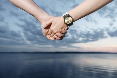 Sweet couple. Close up photo of hands of man and woman holding each other, beautiful sunset on the background, selective focusの写真素材
