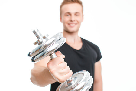 Portrait of a young handsome man wearing black t-shirt standing smiling holding a silver dumbbell in his stretched hand in front of his body, isolated on white background, selective focusの写真素材