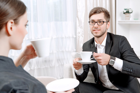 Young man wearing a black suit sitting on a couch drinking tea and talking friendly with his psychologist during therapy session, selective focusの写真素材