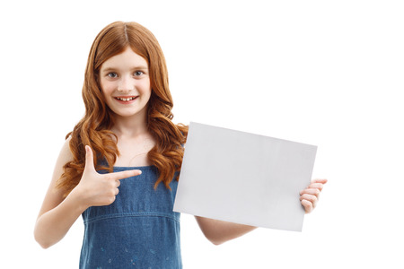 Here it is. Cute red-haired smiling girl holding sheet of paper with one hand and pointing it while showing positivity.の写真素材