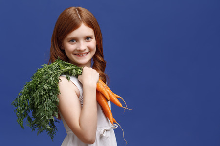 Be healthy. Vivacious red-haired little girl holding the bunch of carrot on her shoulder and smiling while standing half-turned.の写真素材