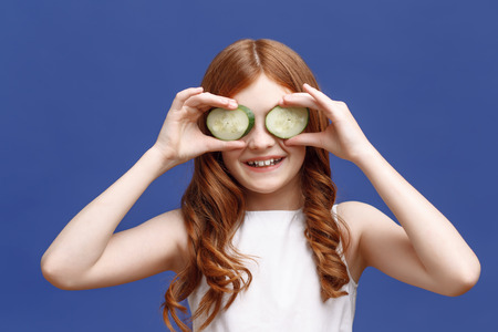 Joyful moment. Upbeat red-haired little girl holding slices of cucumber with two hands on her eyes and smiling while evincing rejoicingの写真素材