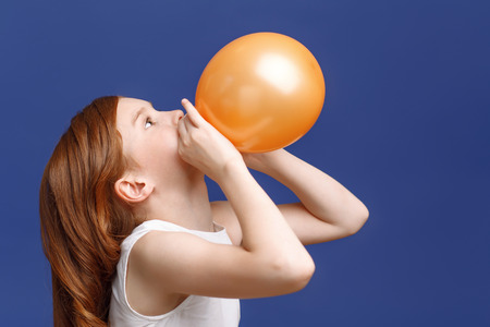 Lets have fun. Close up on pretty young red-haired girl inflating balloon while standing half-turned.の写真素材